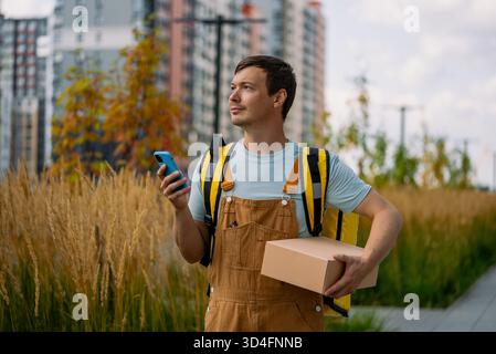 Male delivery driver standing outside in a city, holding a smartphone and a cardboard package, ready to deliver goods to customers, representing freelance work and fast urban shipping Stock Photo