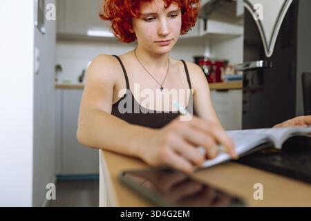 Young red-haired woman sits at table, uses a computer, makes notes in notepad. student studying online, at home Stock Photo