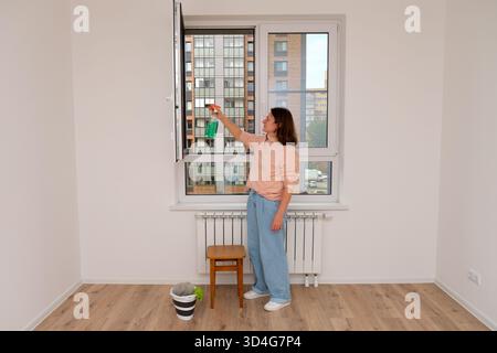 Woman spraying cleaner on an open window, standing on a stool to reach. Spring cleaning concept for apartment management or home maintenance. Stock Photo