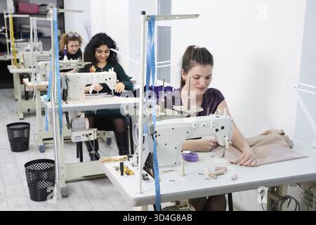 Young dressmaker woman sews clothes on sewing machine. Smiling seamstress and her hand close up in workshop. Focus on sewing machine and tissue Stock Photo
