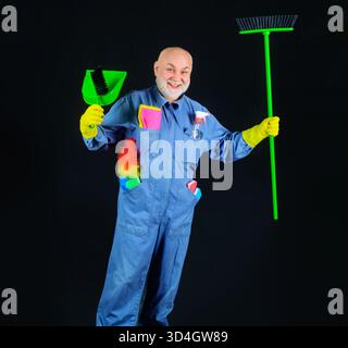Cleaning time. Smiling cleaner with sweeping tools and cleaning supplies. Cleaning agency. Senior man in uniform with broom, dustpan and brush for Stock Photo