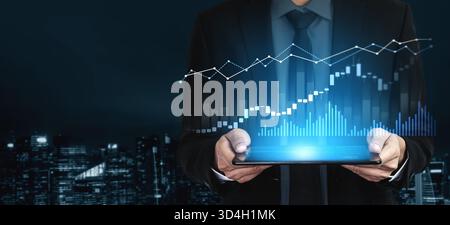 A businessman in a suit holds a tablet displaying vibrant financial graphs and data visualizations against a night city backdrop, symbolizing economic Stock Photo