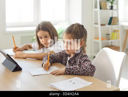 Two children sitting at table and doing homework while using tablet and writing with pencils at home Stock Photo