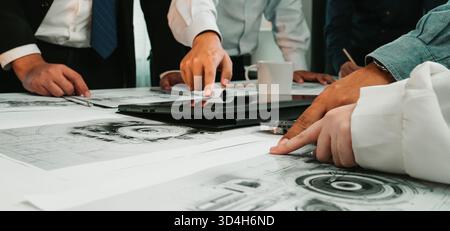 A diverse group of professionals engaged in a business meeting, analyzing technical drawings and discussing plans around a workspace table with Stock Photo