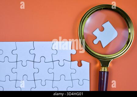 Magnifying glass over white puzzle highlights missing piece on orange background, symbolizing focused search and nearing completion of structured task Stock Photo