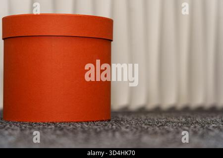 A vibrant orange round box is resting on a textured dark surface, surrounded by a softly lit background. The setting is indoor and cozy. Stock Photo