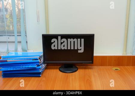A clean workstation features a black monitor on a wooden desk with neatly stacked blue folders nearby. Natural light from the window adds brightness. Stock Photo