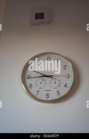 A round clock with gold rim, black numerals, and temp and humidity dials hangs on a light wall under a small square vent in a bright modern apartment Stock Photo