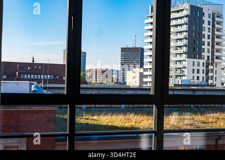 Late afternoon view through a large apartment window in Gdansk shows glass high rises, an old red brick rooftop with dormers and spire, rail lines, an Stock Photo
