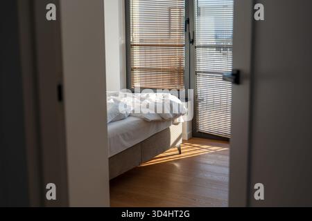 Soft morning light through Venetian blinds falls on wood floors and a low platform bed in a modern Gdansk apartment, viewed from a doorway, suggesting Stock Photo