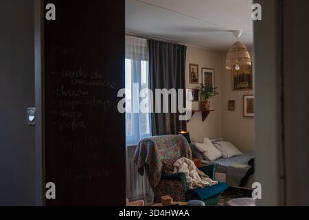 Soft afternoon light filters through sheer curtains and gray drapes. A teal armchair, daybed, rattan lamp, and chalkboard wall with Polish notes set a Stock Photo