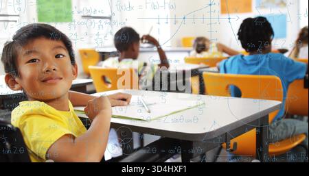 Sitting boy wearing yellow T-shirt writing in workbook at elementary classroom, with math overlay Stock Photo