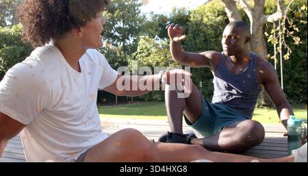 Sitting two men in activewear chatting and gesturing on wooden deck in backyard, with green bottle Stock Photo