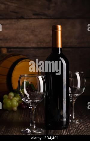 A dark bottle of wine sits next to two empty glasses on a wooden table. Fresh grapes in various colors are nearby, and a barrel is faintly visible in Stock Photo