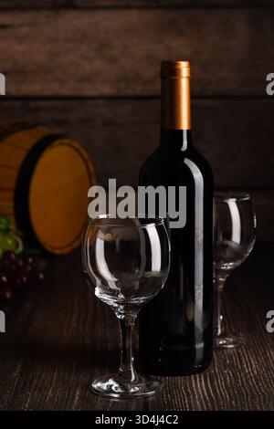 A red wine bottle stands beside two empty glasses on a dark wooden table. Grapes lie nearby, and a rustic barrel adds charm to the scene, suggesting a Stock Photo