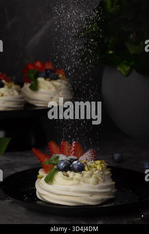 Delicate meringue desserts are beautifully arranged on a dark plate, adorned with fresh strawberries, blueberries, and mint. A sprinkle of sugar adds Stock Photo