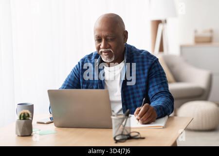 Senior Black Man Working at Home Using a Laptop and Taking Notes While Learning Online Stock Photo