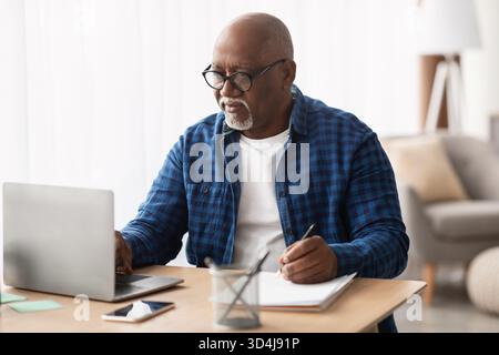 Senior Black Man Focused on Laptop While Taking Notes at His Office Desk Stock Photo