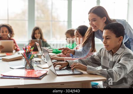 Teacher Guides Diverse Students in Classroom Using Laptop for Online Learning Stock Photo