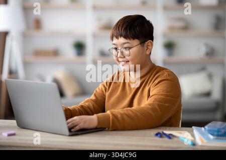 Smart Asian Boy Doing Homework at Home During Pandemic With Modern Laptop and Stationery Stock Photo