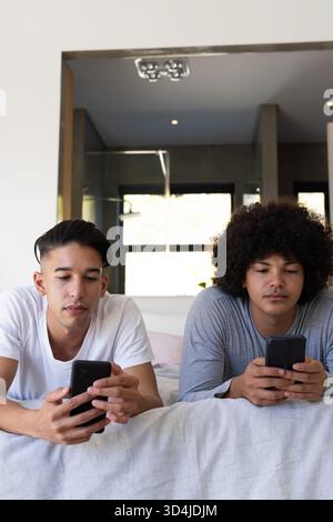 Lying diverse male friends using smartphones on duvet in modern bedroom, showing relaxed routine Stock Photo