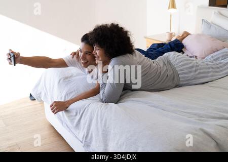 Smiling Diverse male couple leaning on bed in bedroom at home, with smartphone and pastel pillows Stock Photo