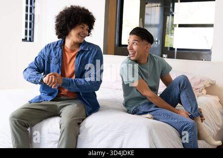 Smiling and laughing Diverse male friends sitting on bed with white pillows in bright bedroom Stock Photo
