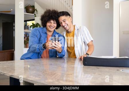 Leaning diverse male friends looking at smartphone on marble island in kitchen, with smartwatches Stock Photo