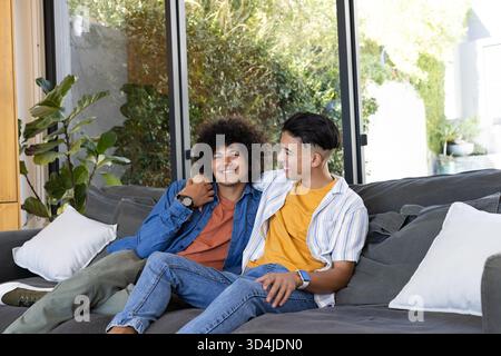 Sitting diverse male couple cuddling closely on gray sofa in living room, with sliding glass doors Stock Photo