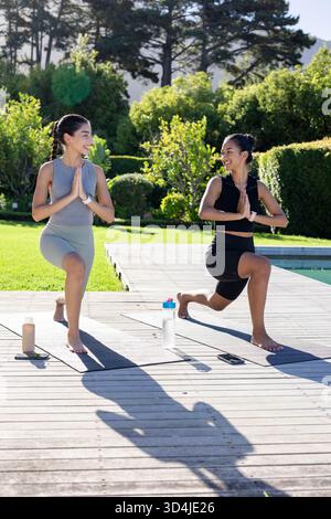 Practicing Diverse female friends doing yoga lunges on poolside deck, with yoga mats, water bottles Stock Photo