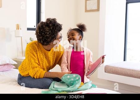 Smiling diverse mother and daughter packing school supplies on bed, with backpack and tablet Stock Photo