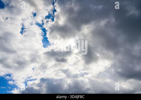 A horizontal view of a dramatic sky, with the sun breaking through a mix of white, fluffy, and dark, stormy clouds, revealing patches of blue. Stock Photo
