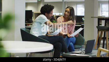 Discussing two colleagues reviewing documents in open-plan office lounge, laptop and glass on table Stock Photo
