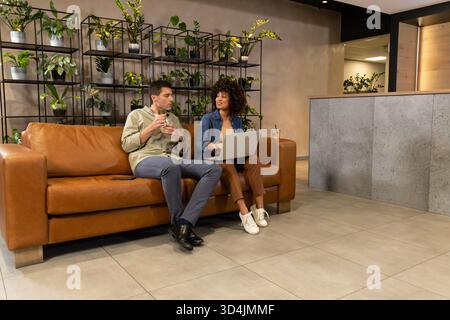 Sitting Diverse coworkers chatting on leather sofa in office lounge, with laptop and coffee cup Stock Photo
