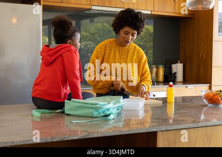 Diverse mother and daughter chopping carrots on cutting board in kitchen, with mint green lunchbox Stock Photo