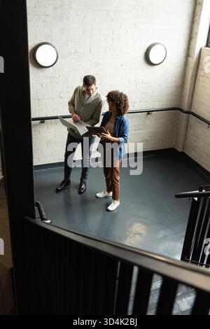 Standing diverse coworkers reviewing documents on stairwell landing, with silver laptop and folder Stock Photo