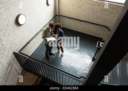 Discussing Diverse coworkers reviewing documents on stairwell landing, with tablet and blueprints Stock Photo