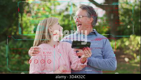 Laughing bespectacled man and ringed woman holding smartphone in sunlit garden with HUD, sweaters Stock Photo