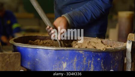 Mixing worker's hands in blue shirt pushing rod into brown mixture at workshop, with blue tub Stock Photo