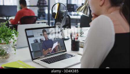 Watching remote presenter wearing dark top holding papers on laptop at office desk, lamp Stock Photo
