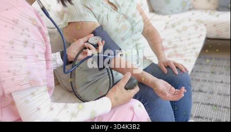 Measuring nurse in pink uniform wrapping navy pressure cuff at home, holding gauge and stethoscope Stock Photo