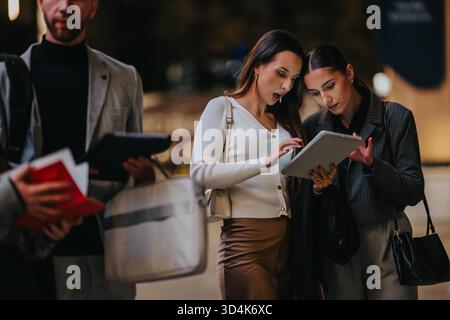 Two businesswomen review tablet with colleague in urban night scene, collaborative teamwork Stock Photo