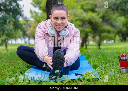 Sporty woman stretching leg sitting on yoga mat outdoors in park smiling Stock Photo