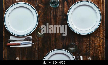 A top-down view of a rustic wooden table set with three white oval plates, silverware, and napkins, ready for a meal. Stock Photo