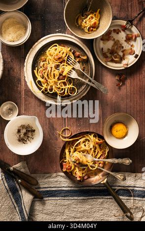 top view of wooden table set with bucatini carbonara in copper pan and plates and bowls accompanied with egg yolk, bacon and pepper in mortar, rustic Stock Photo