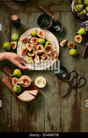 flay lay of ripe figs in plate and in a basket ready to eat with goat cheese and thyme honey,someone is holding the plate Stock Photo