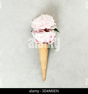 Whimsical composition of pink peonies arranged in a waffle cone, resembling ice cream, placed on a light gray textured background. Stock Photo