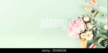 Creative arrangement of pastel flowers and colorful macarons in a waffle cone, styled on a mint green background with space for text. Stock Photo
