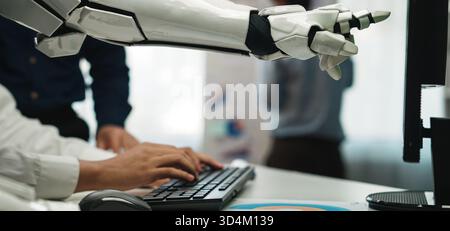 A robotic arm assists a human worker at a keyboard in a modern office, showcasing innovative technology and collaboration in a digital workspace Stock Photo