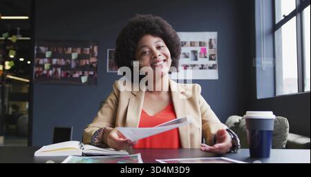 Smiling African American woman in beige blazer handing printed pages at office table with notebook Stock Photo